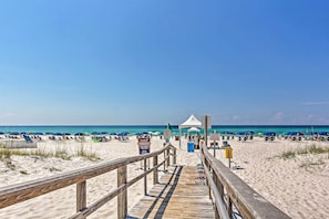 Ein hölzerner Steg führt zu einem Strand mit Sonnenschirmen und Liegestühlen.