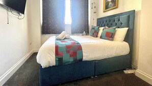 Un letto rifatto con cura, con una testiera blu, una coperta e un asciugamano arrotolato sopra. Al muro, sopra il letto, c'è un quadro.