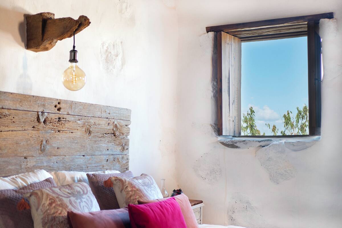Une chambre à coucher au style rustique, avec une tête de lit en bois, un lit recouvert d’une literie à motifs, une petite table de chevet et une fenêtre donnant sur les arbres.