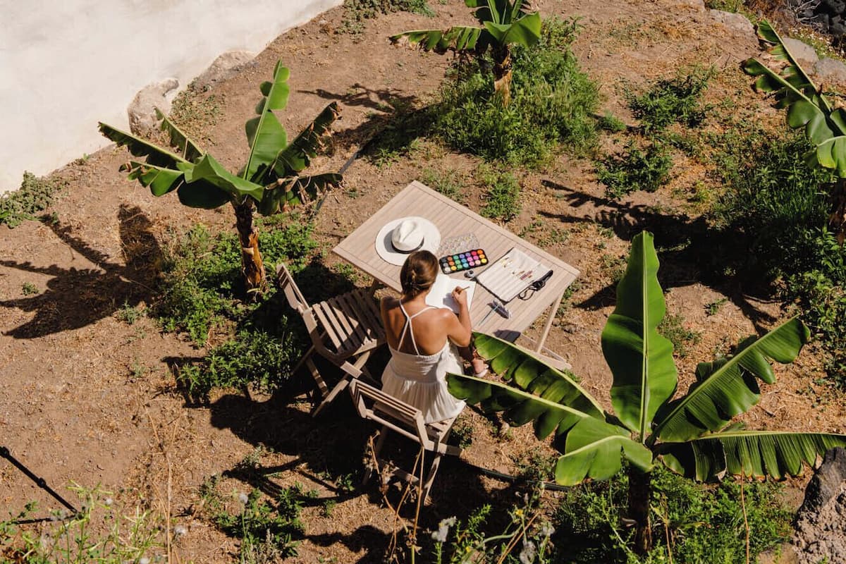 Une scène côtière avec un bâtiment blanc, une table sur laquelle se trouvent un chapeau et une palette, et une femme qui peint.
