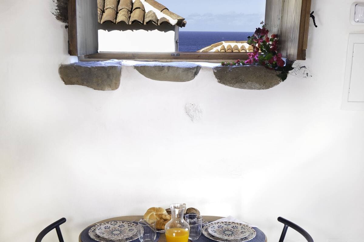 Une table à manger avec deux chaises, une fenêtre donnant sur la mer et un petit arrangement floral.