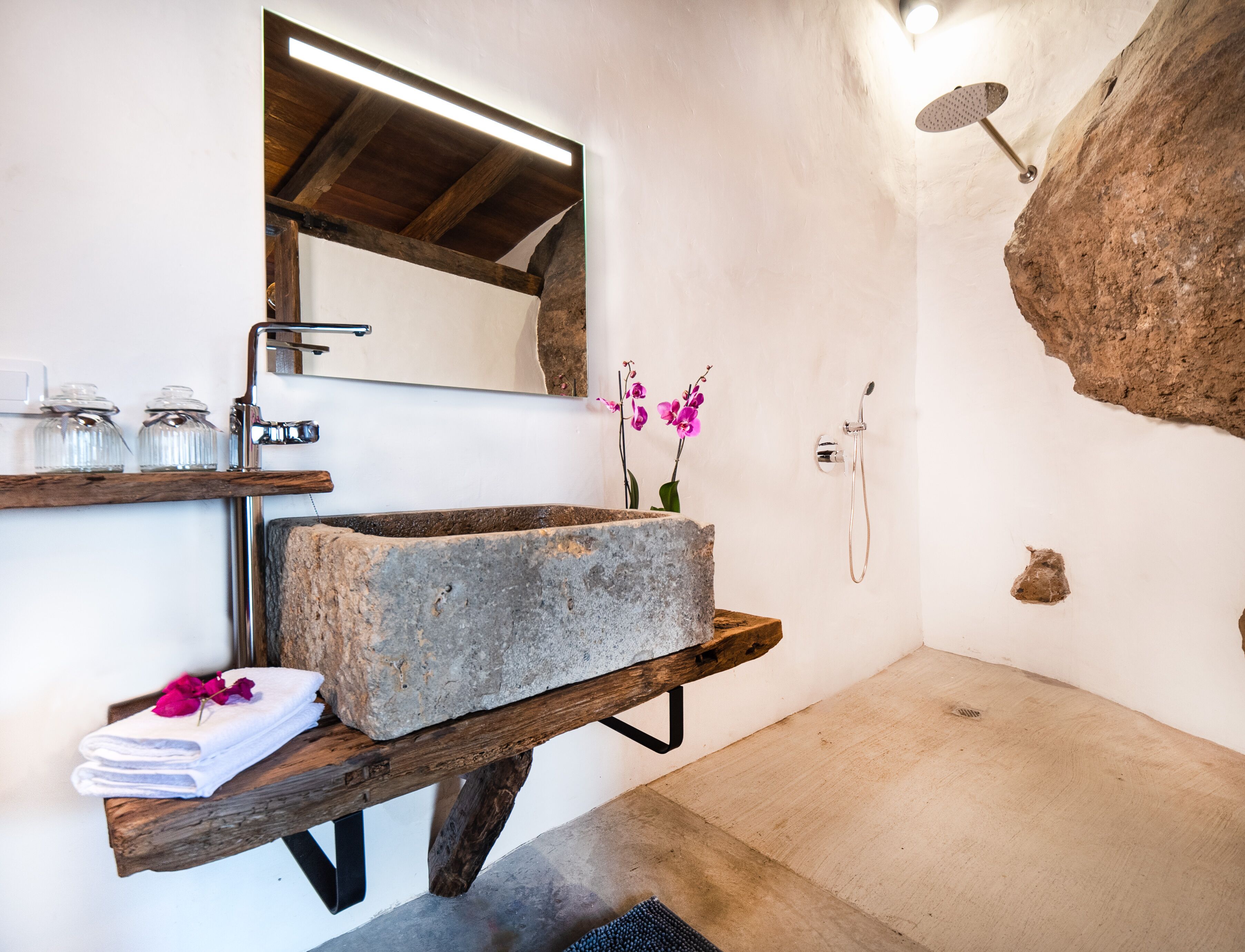 A bathroom with a stone sink, wooden shelf, and a shower area.