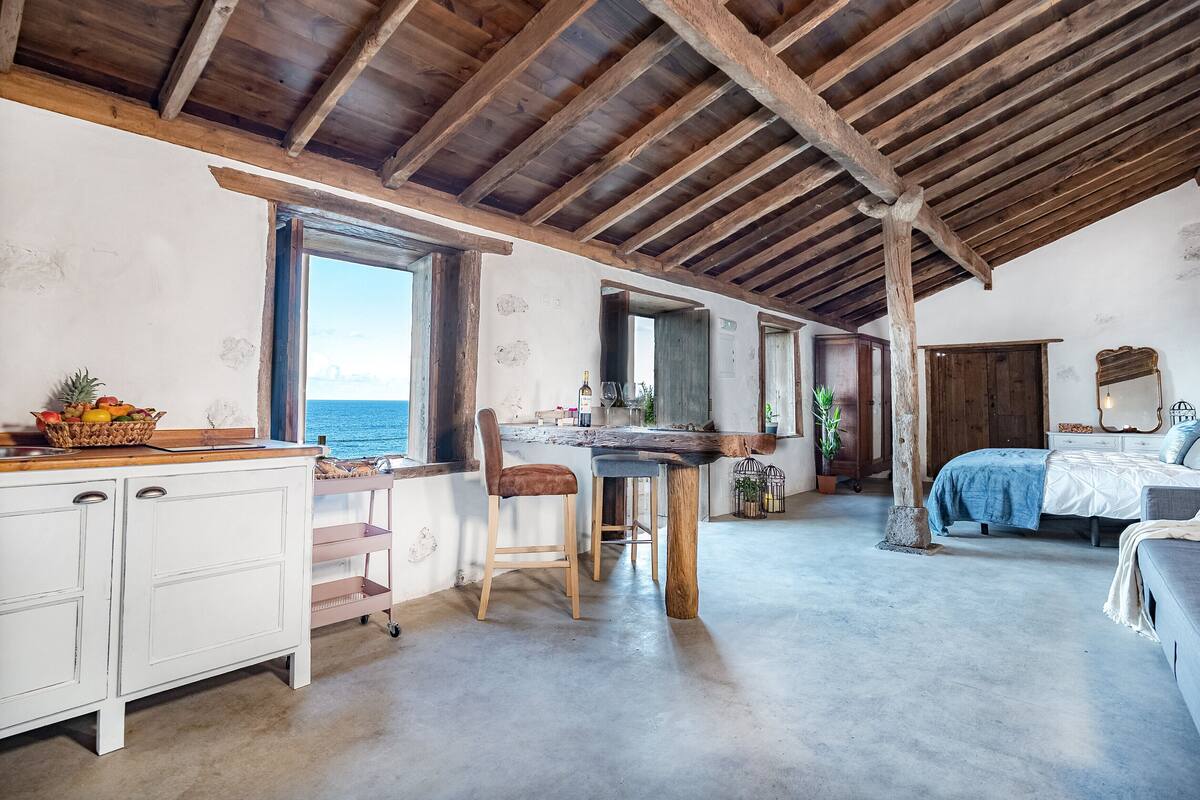 Une pièce rustique avec un plafond en bois, un coin repas avec une table et des chaises, un lit recouvert d’une couverture bleue, et une vue sur la mer par la fenêtre.