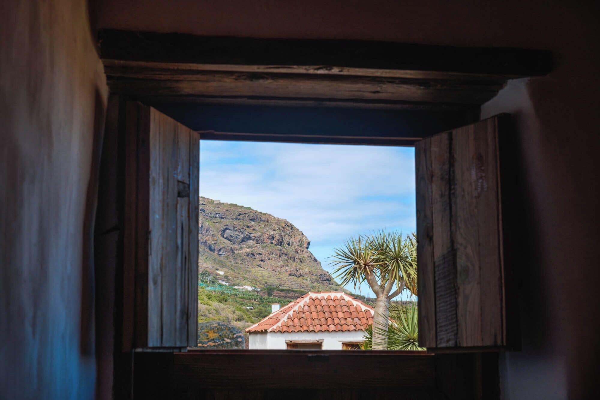 A view through a window with wooden frames, a mountain in the background, and a building with a red-tiled roof.