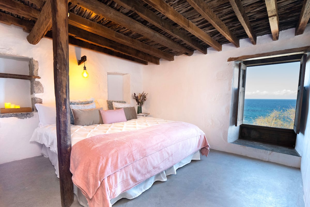 Une chambre à coucher avec un lit, une table de chevet, une fenêtre donnant sur la mer et un plafond en bois.