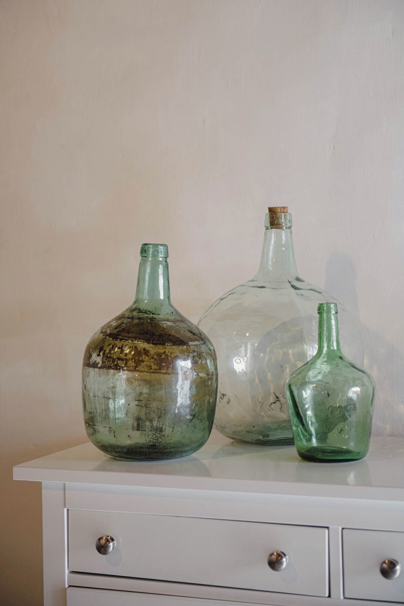 Three glass bottles of varying sizes on a white dresser against a plain wall.