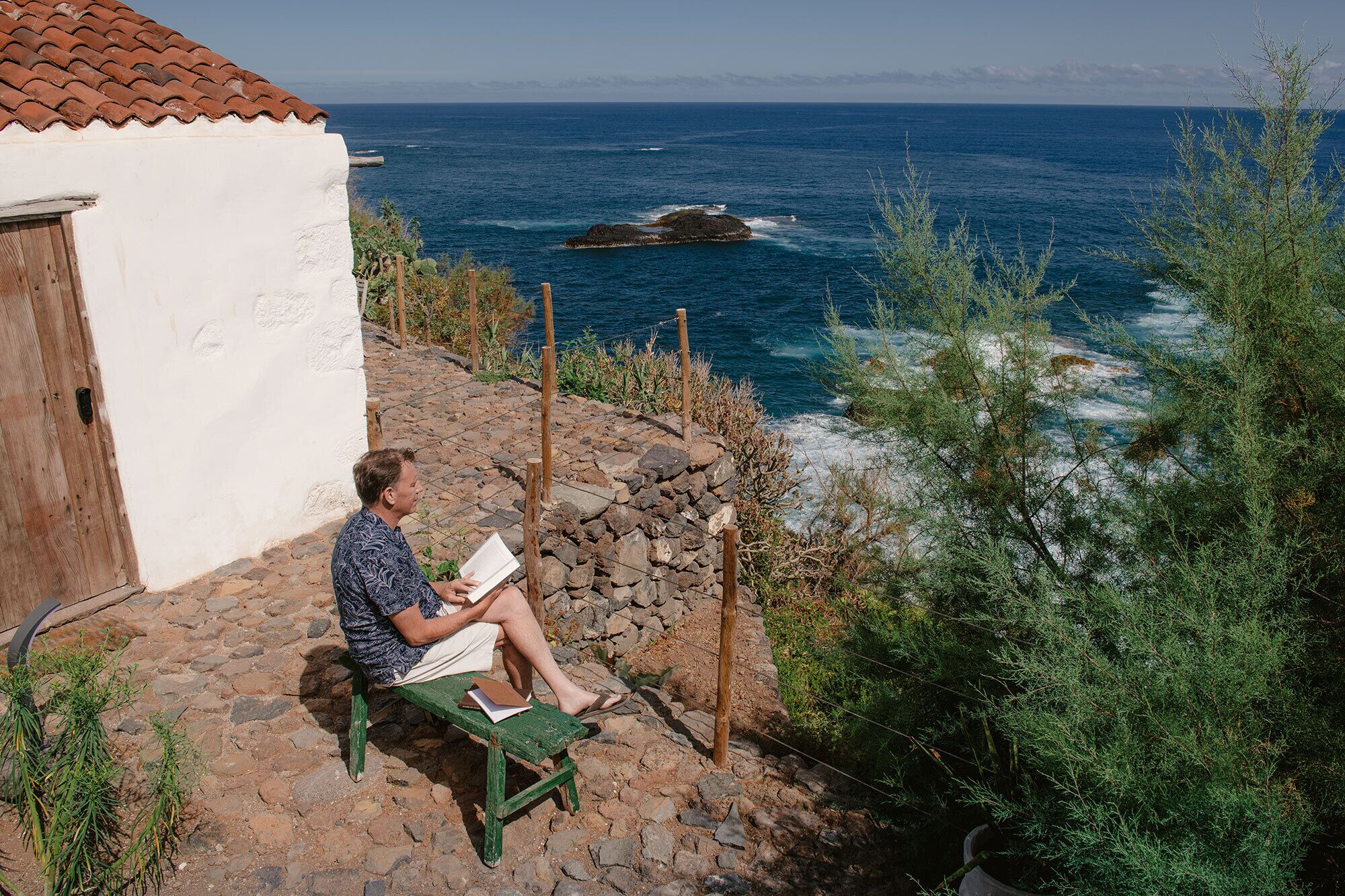 Un hombre leyendo un libro en un banco de piedra junto a un edificio costero, con vista al mar.