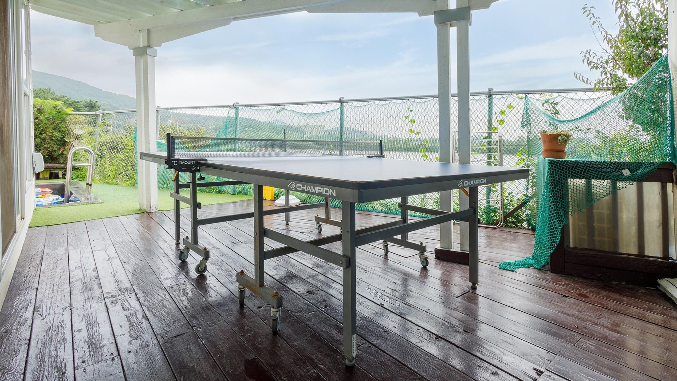 Mesa de ping pong en una terraza de madera con vistas a un área verde con red y un jardín.