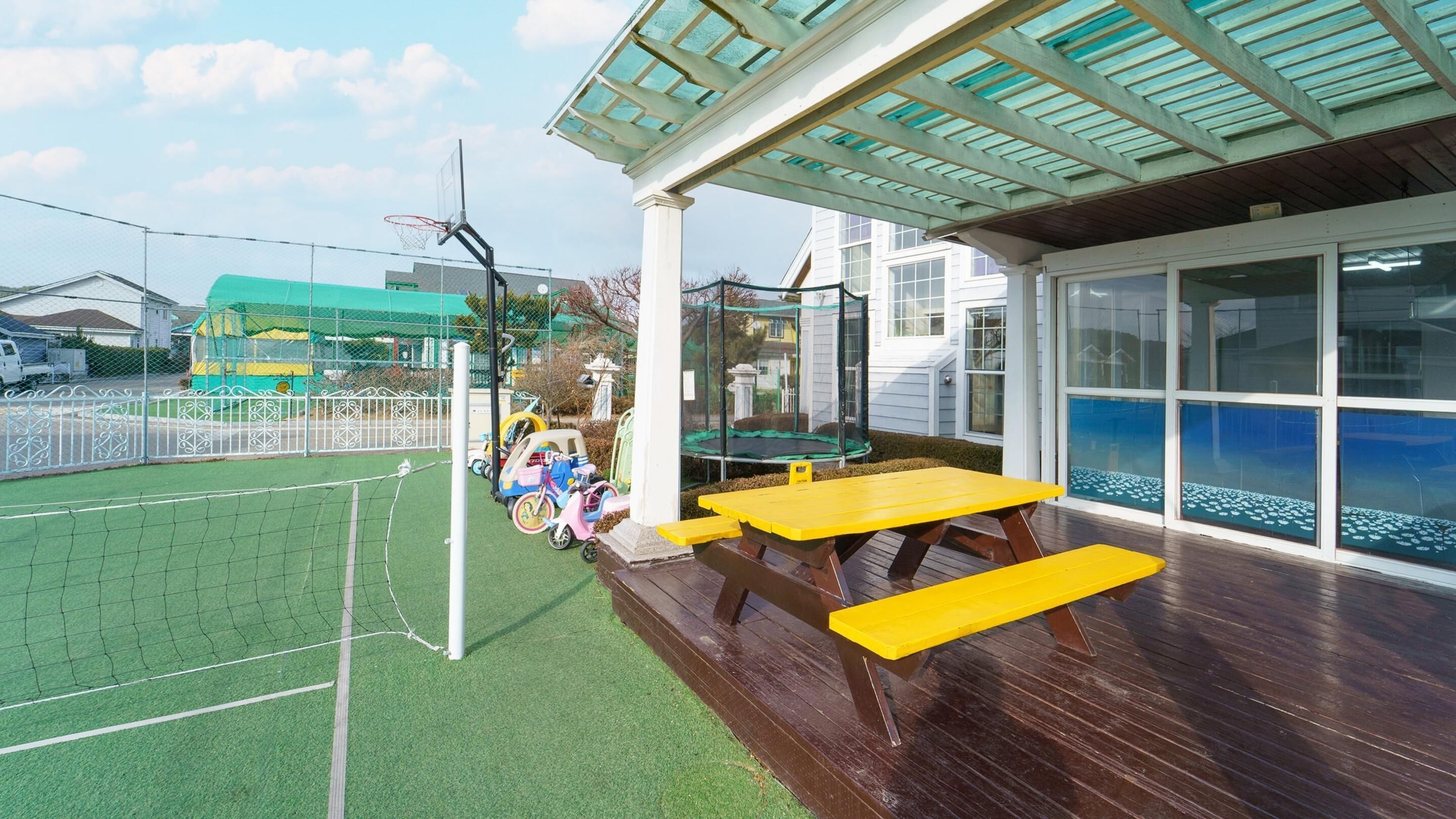 A covered outdoor area with a green artificial turf sports court, a yellow picnic table, and a transparent enclosure for a trampoline.