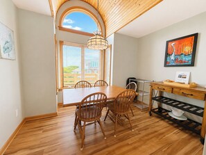 Un coin repas avec une table et des chaises en bois, un luminaire suspendu, une vue par la fenêtre et un tableau accroché au mur.