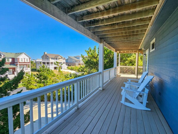 A wooden porch with white railings and rocking chairs, overlooking a residential area with houses and greenery.