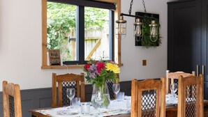 Ein Essbereich mit einem Holztisch, gedeckt für sechs Personen, umgeben von Stühlen mit Gitterrücken. Auf dem Tisch steht eine Vase mit Blumen, darüber hängen Leuchten.