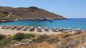 Une plage avec des chaises longues et des parasols en chaume, une mer d’un bleu limpide et, en arrière-plan, une colline rocheuse.