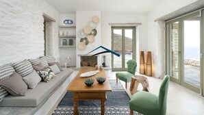 Un salon moderne avec un mur en pierre, une table en bois, un fauteuil vert et une vue sur la mer grâce à de grandes portes vitrées.