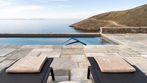 Une piscine en pierre, dotée de chaises longues et de serviettes, qui offre une vue sur une mer calme et un paysage vallonné.