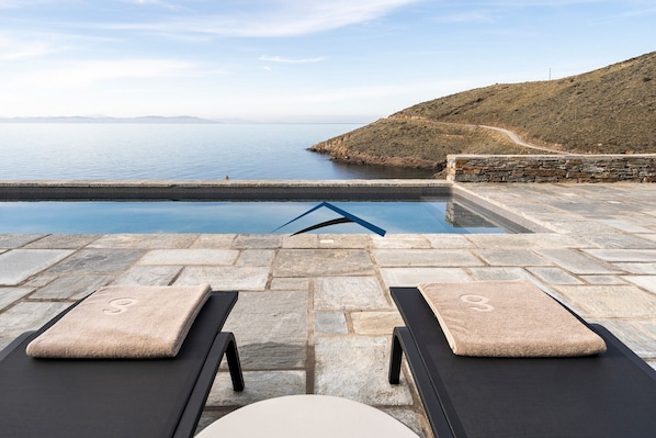 A stone pool with lounge chairs and towels, overlooking a calm sea and a hilly landscape.