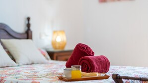 Un lit bien fait, recouvert d’une couvre-lit à motifs floraux, une paire de serviettes rouges, un verre de jus d’orange et un plateau en bois avec un bol et une cuillère.