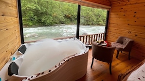 A wooden cabin with a large window overlooking a river, a hot tub, and a chair. - Teona Caglayan Bungalov (Findikli)