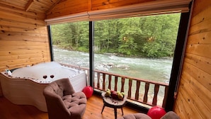 A wooden cabin with a hot tub, two chairs, a small table with fruit, and a view of a river. - Teona Caglayan Bungalov (Findikli)