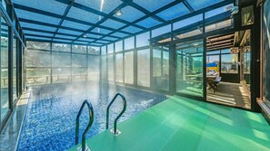 An indoor swimming pool with clear glass walls, a green floor, and a visible outdoor area with a blue fence. - Daebudo Juni Pension (Ansan)