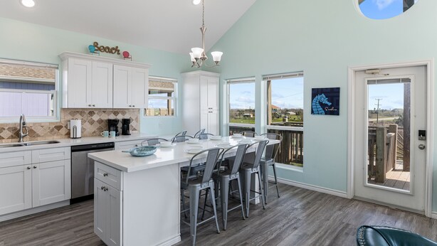 A modern kitchen with white cabinetry, a central island with bar stools, and large windows offering views of the outdoors. - Steps from the Beach!  (Crystal Beach)