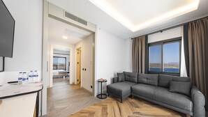 Une chambre d’hôtel moderne avec un canapé gris, un plancher en bois, une petite table ronde et une grande fenêtre donnant sur un beau paysage.