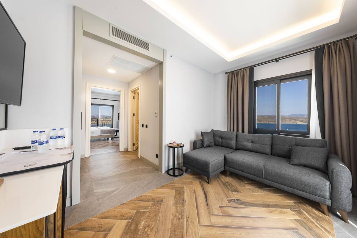 A modern hotel room with a grey sofa, wooden flooring, a small round table, and a large window with a view.