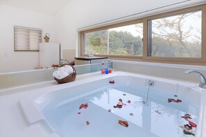 A modern bathroom with a large bathtub, a window with a view of trees, and a basket of towels.