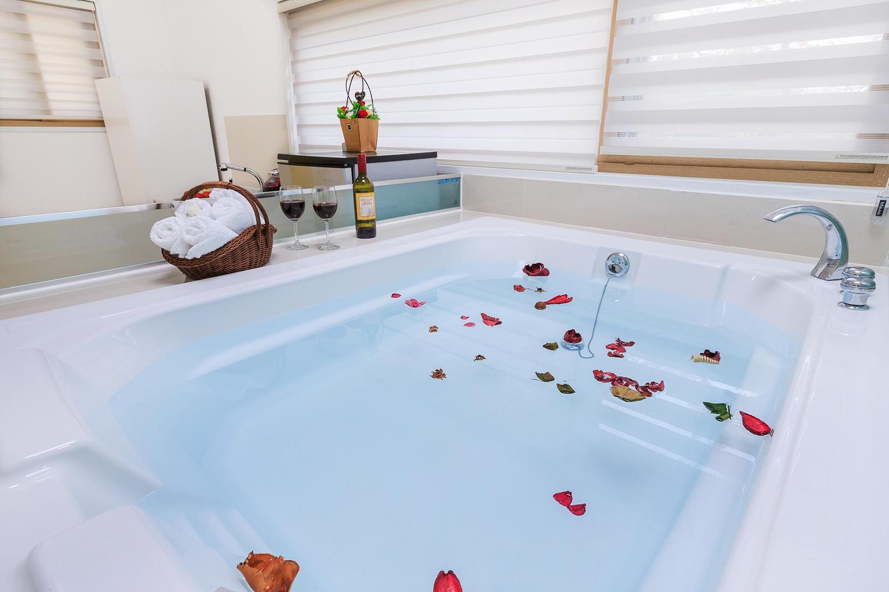A bathtub with rose petals and a bottle of wine, a basket of towels, and a wine glass.