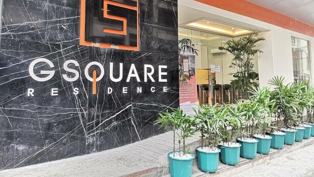 Una pared de mármol negro con el texto "GSQUARE RESIDENCE" y un logotipo. Hay plantas en macetas al frente.