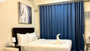 Habitación de hotel con cama, mesita de noche, lámpara, silla, mesa de vidrio y cortina azul.