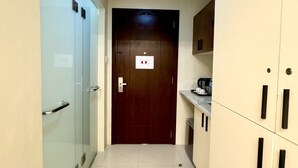 Un pasillo estrecho con una puerta de madera al final, una puerta de cristal a la izquierda y una zona de kitchenette a la derecha.