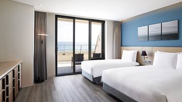 A hotel room with two beds, a view of the sea, a balcony, and a desk with storage.