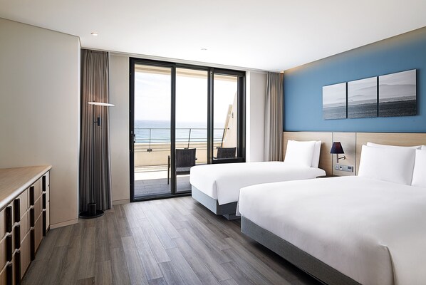 A hotel room with two beds, a view of the sea, a balcony, and a desk with storage. - Parnas Hotel Jeju (Seogwipo)