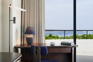 A modern hotel room with a desk, a lamp, a chair, a cup, and a book.