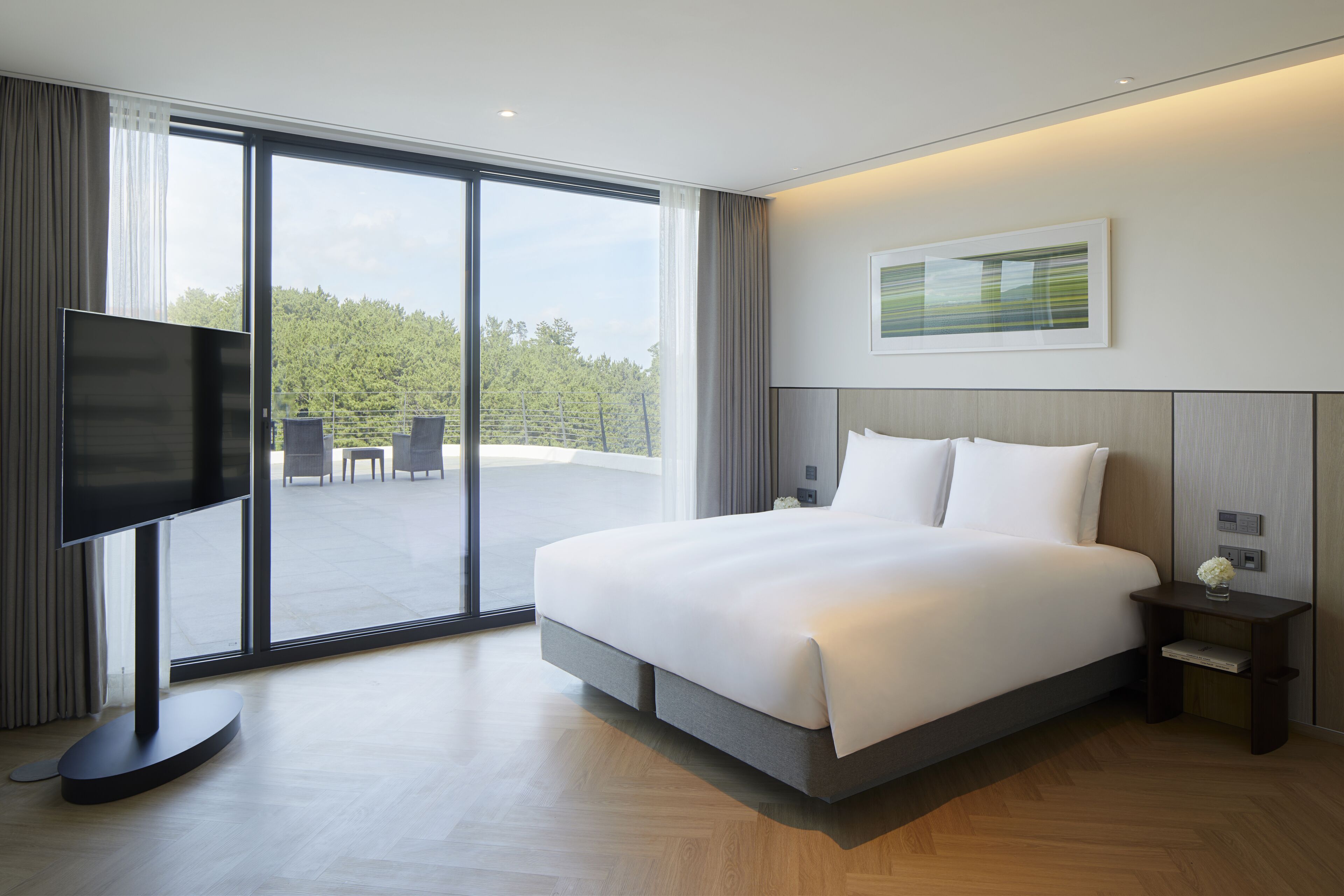 Habitación de hotel moderna con una cama grande, un televisor de pantalla plana, una mesita con un jarrón y vistas al exterior a través de amplias ventanas.