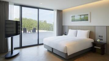 A modern hotel room with a large bed, a flat-screen TV, a small table with a vase, and a view of the outdoors through large windows.