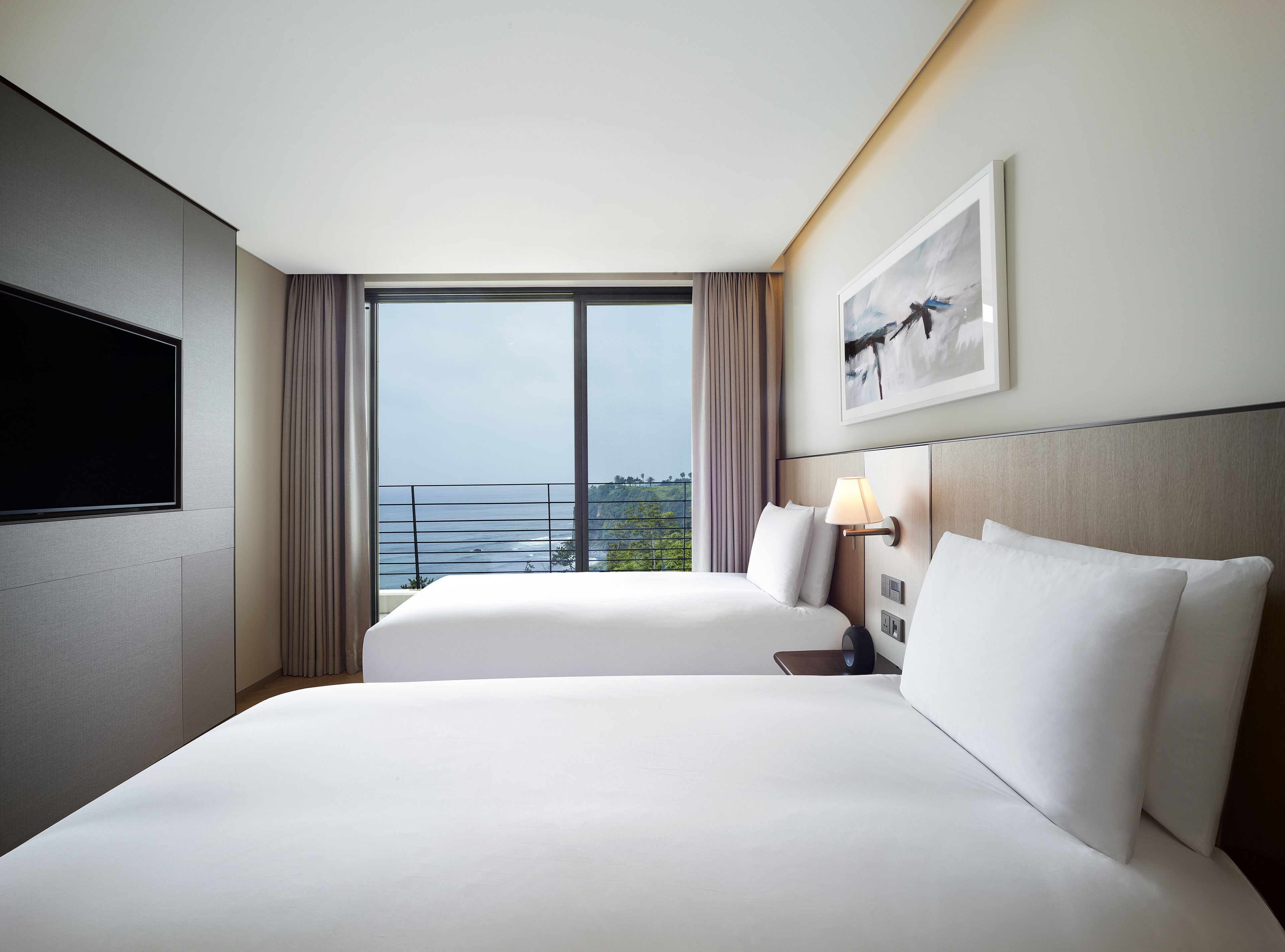 A hotel room with two beds, a TV, a painting, and a view of the ocean.