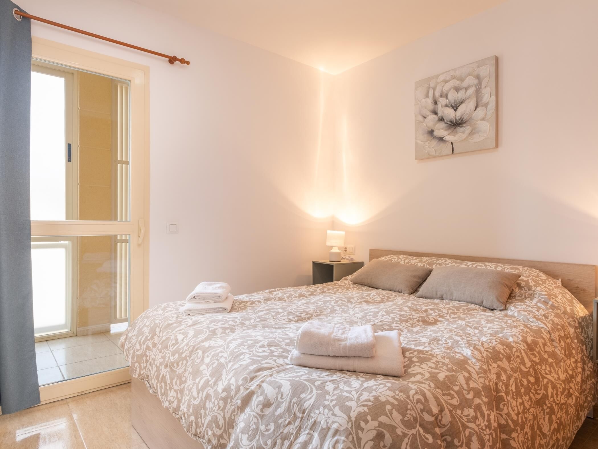Une chambre avec un grand lit, une table de chevet avec une lampe, une fenêtre avec des rideaux et une œuvre d’art florale accrochée au mur.
