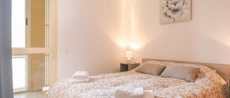 Une chambre avec un grand lit, une table de chevet avec une lampe, une fenêtre avec des rideaux et une œuvre d’art florale accrochée au mur.