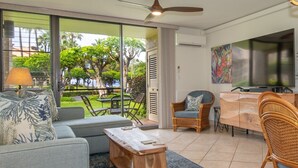 A modern living room with a sofa, a wooden coffee table, wicker chairs, a dining area with a table and chairs, and a large TV. - Maui Paradise: Steps to Sand-Blg#1. Partial Ocean View, AC's, Wifi, 1st Floor (Kihei)