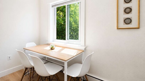 A room with a dining table, two chairs, a window with a view of greenery, and a framed picture on the wall.