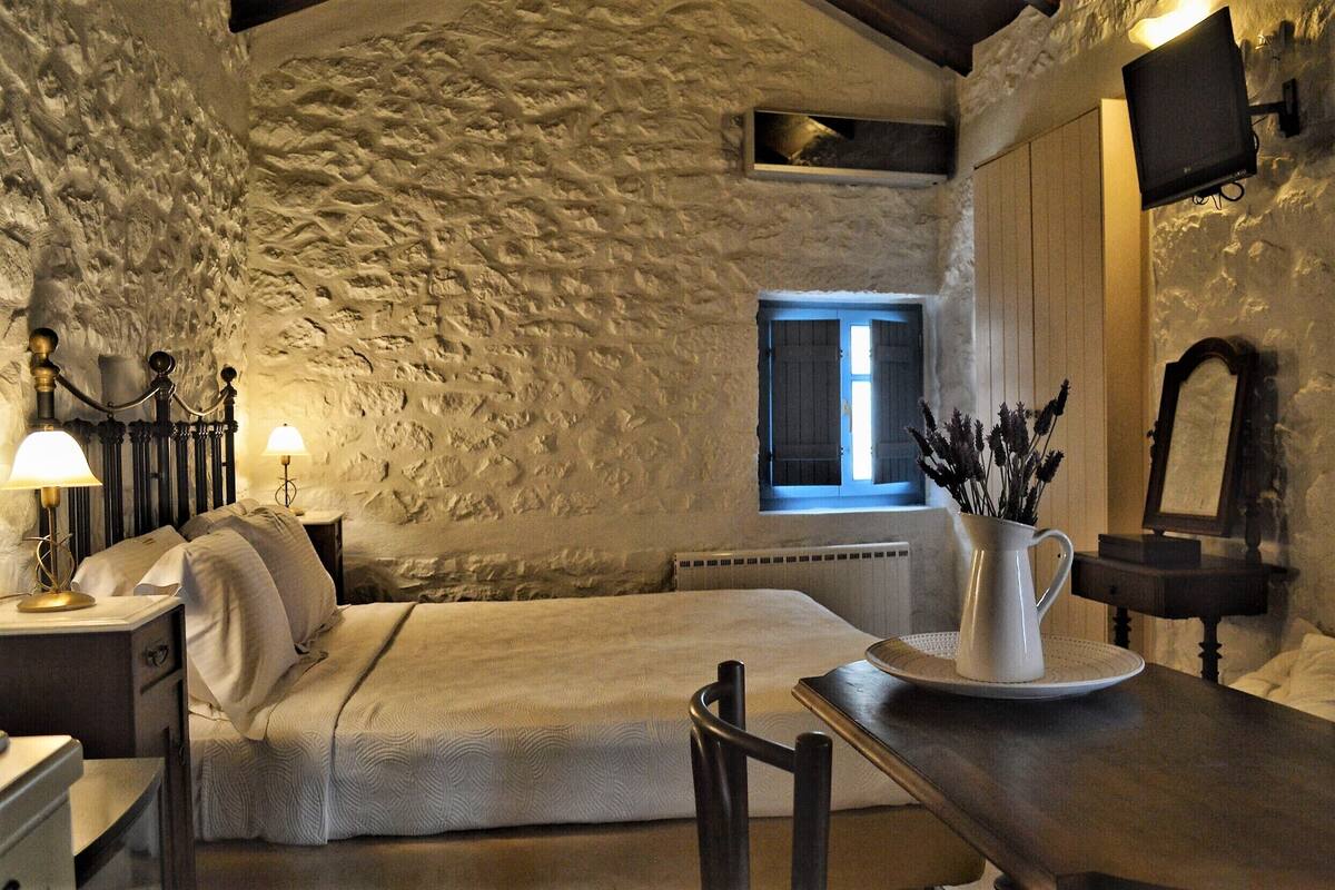 A cozy bedroom with a stone wall, a bed, a nightstand with a lamp, a dresser, a table with a pitcher, and a window with curtains.