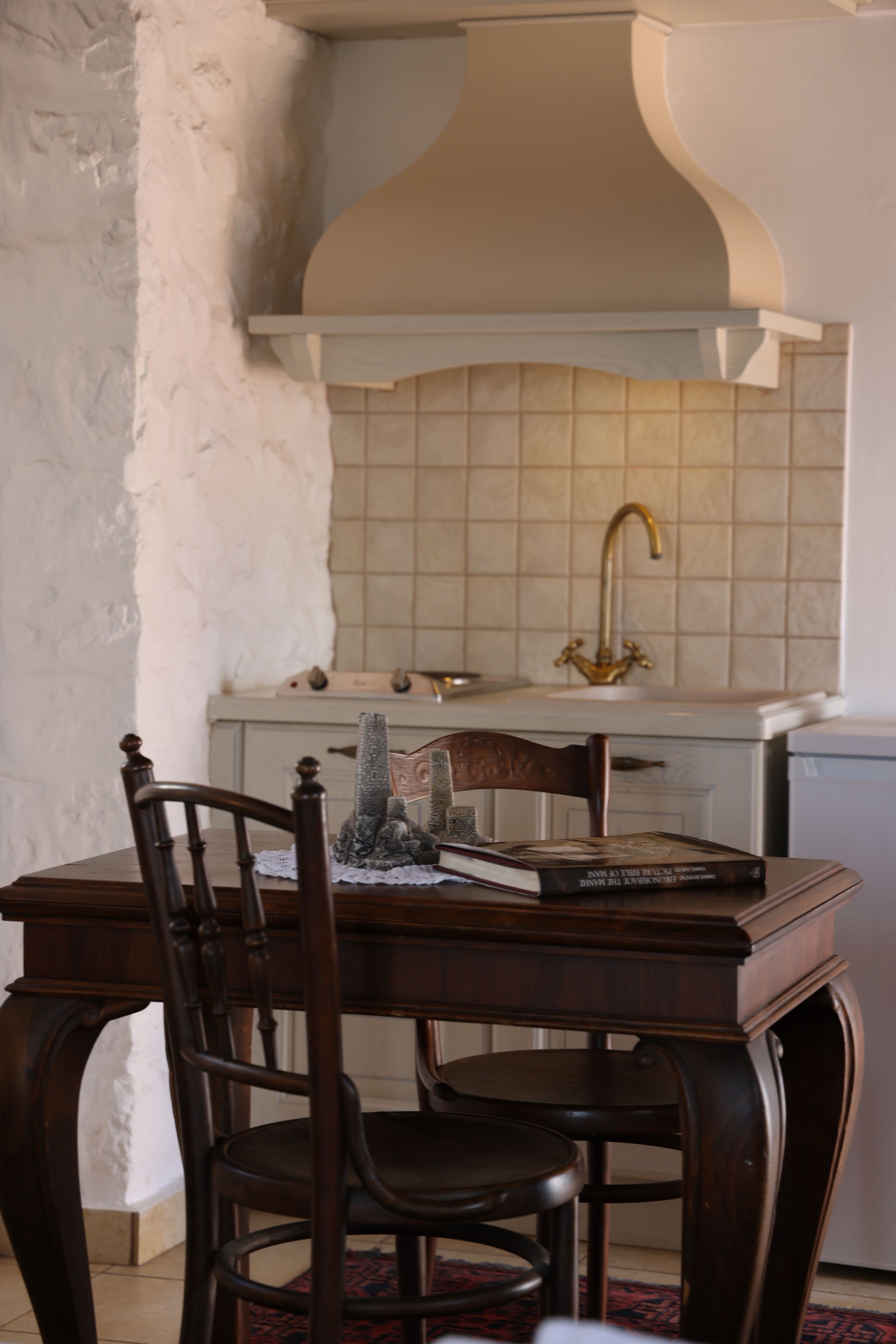 A dining area with a wooden table and chairs, a decorative book, and a kitchen in the background with a tiled backsplash and a brass faucet.