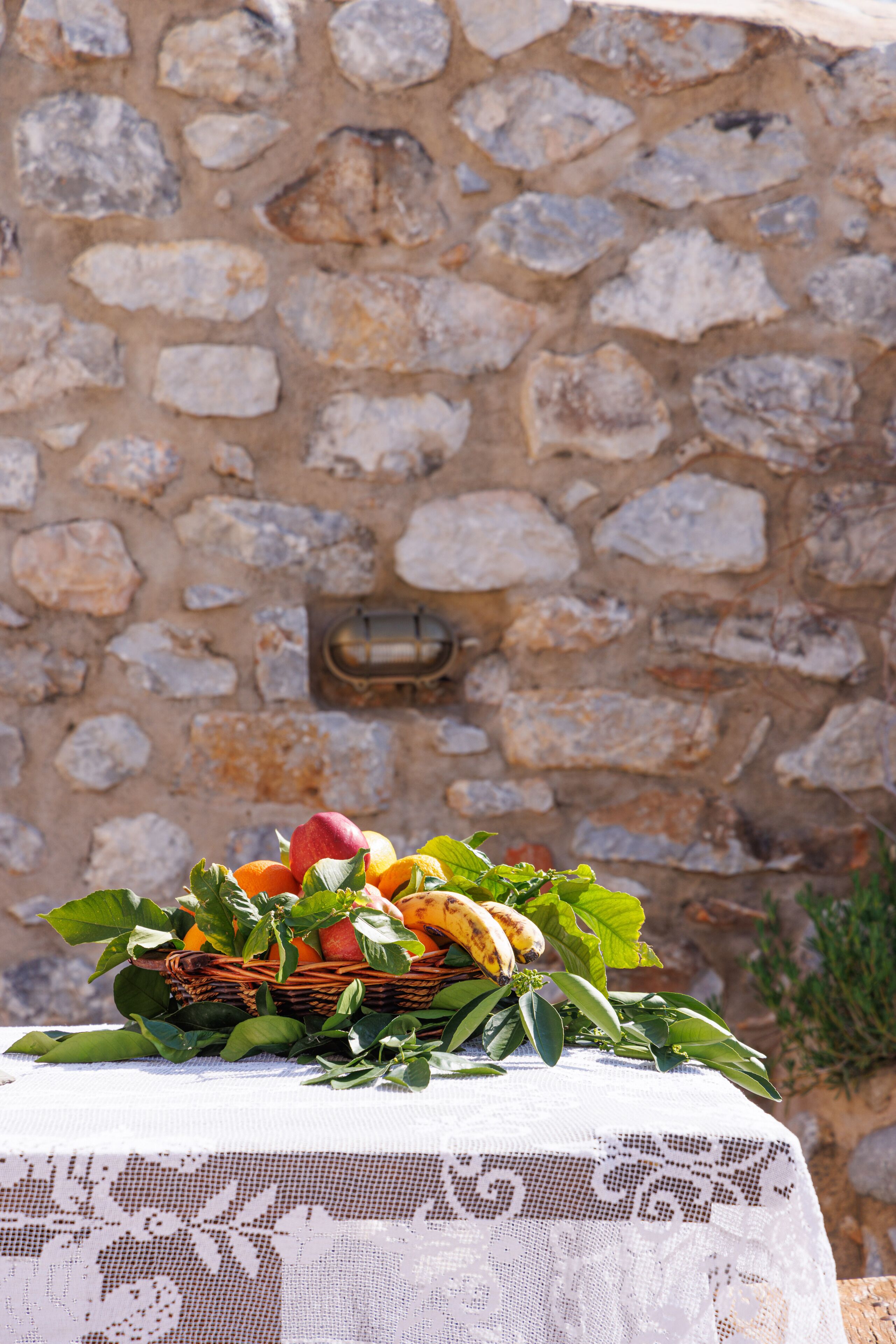 Una mesa con mantel de encaje, una cesta de mimbre llena de frutas y hojas, y un muro de piedra de fondo.