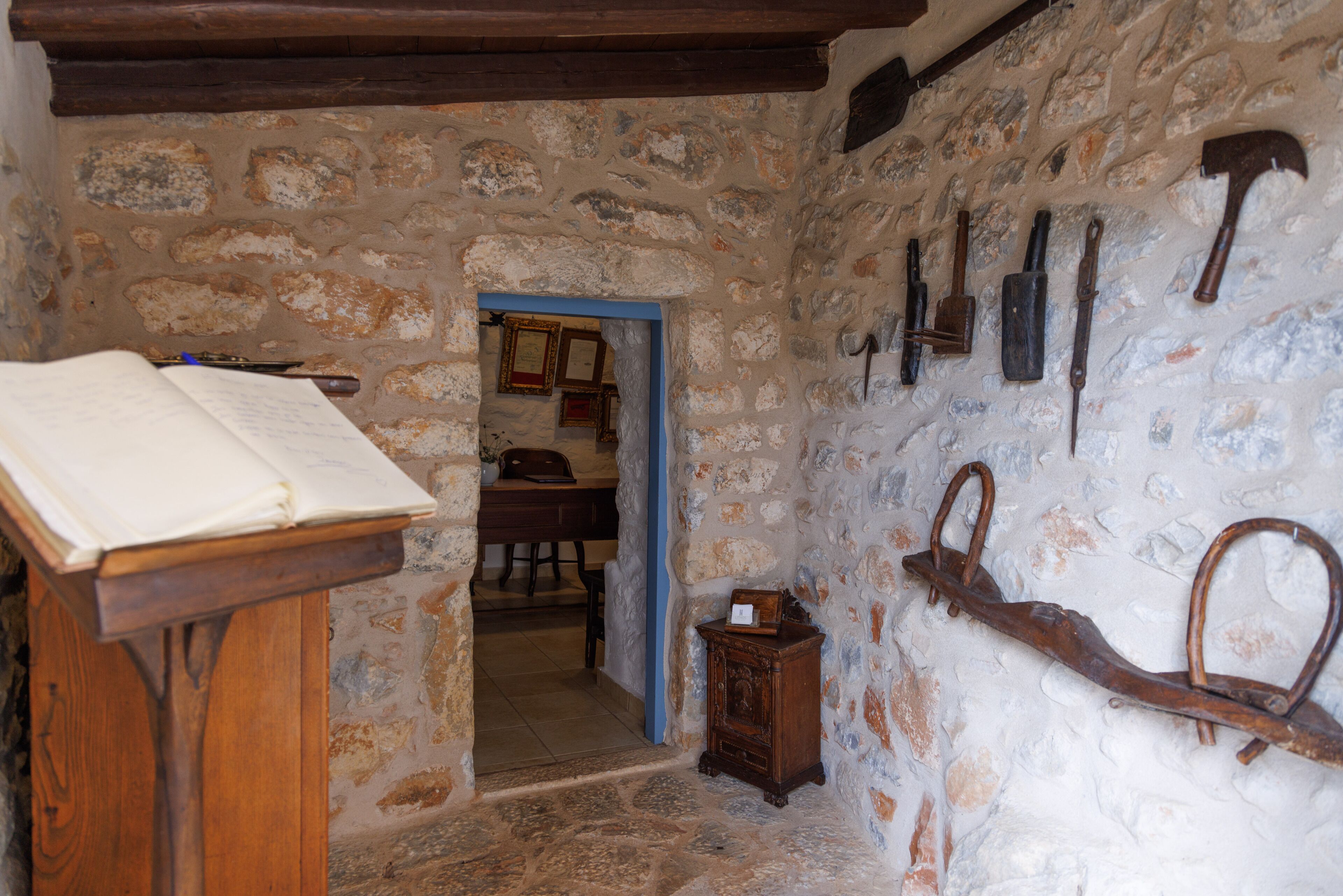 Une pièce rustique avec un mur en pierre, un bureau en bois sur lequel repose un livre ouvert, et divers outils accrochés au mur.