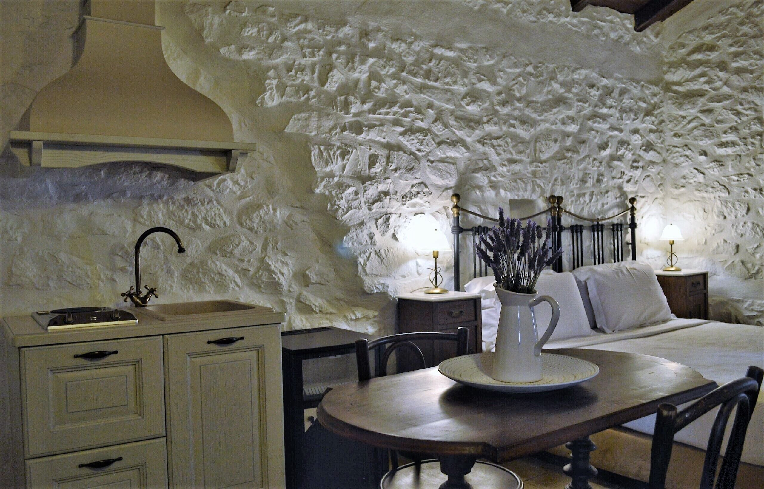 A rustic room with a stone wall, a wooden table, a white pitcher, and a bed with white bedding.