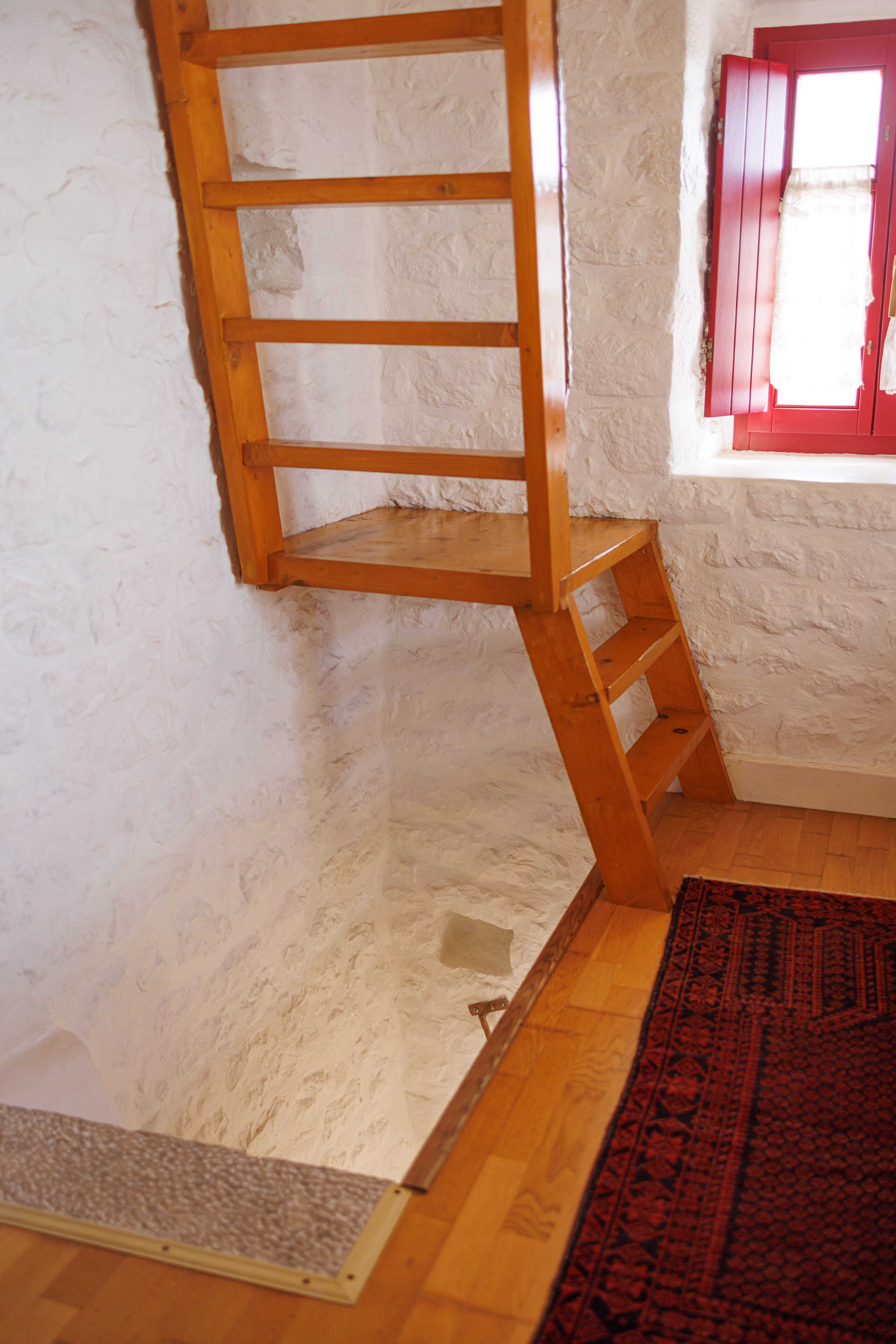 A room with a wooden ladder shelf, a red window with curtains, and a patterned rug on the floor.