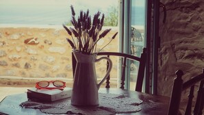 Una mesa con un jarrón de lavanda, un par de gafas y un libro, frente a una pared de piedra con vista a la playa.