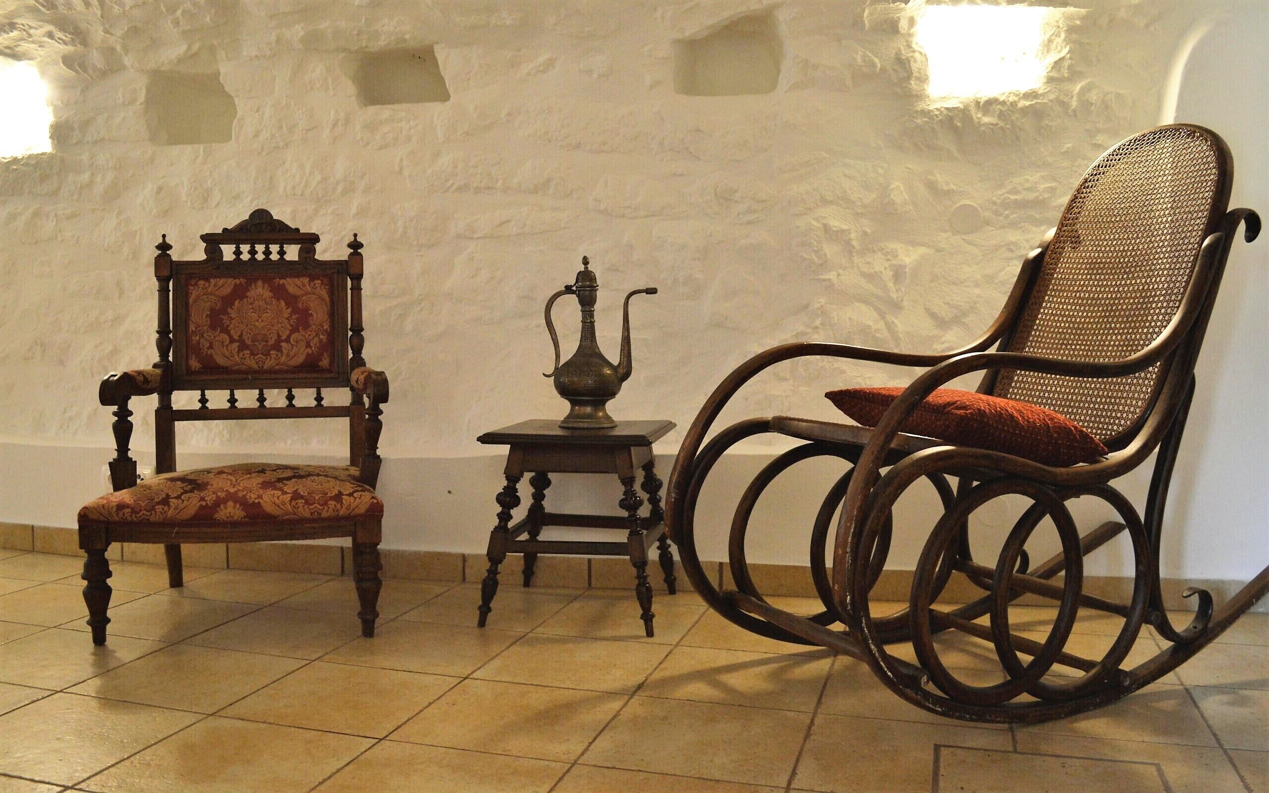 Une pièce comprenant une chaise en bois avec un siège rembourré, un fauteuil à bascule avec un dossier en osier, et une petite table sur laquelle se trouve une théière en métal.